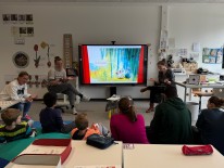 Schüler:innen in Sitzkreis vor einer digitalen Tafel, auf der ein Bilderbuchkino gezeigt wird
